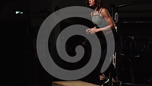 Beautiful female fitness athlete performs box jumps in a dark gym wearing black sports top and short tights with face