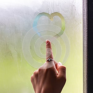 A beautiful female hand draws heart on misty window. finger on the wet glass draws the heart, window and rain