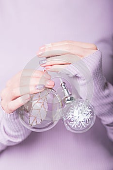 Beautiful female hands holding two transparent Christmas balls on the pale violet background. Manicure with pink color nail polish