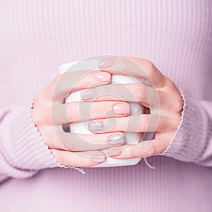 Beautiful female hands holding white cup on the pale violet background. Manicure with pink color nail polish with shiny design.