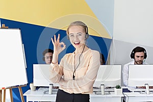 Beautiful female helpline consultant looking at camera and showing OKAY gesture in modern office