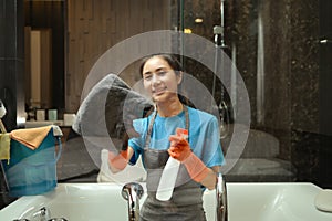 Beautiful female housekeeper in rubber gloves cleaning mirror with rag and spray in a modern bathroom