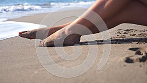 Beautiful female legs lying on sand and relaxing near sea shore. Young woman sitting on the coast and tanning on a sunny