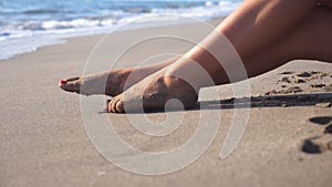 Beautiful female legs lying on sand and relaxing near sea shore. Young woman sitting on the coast and tanning on a sunny