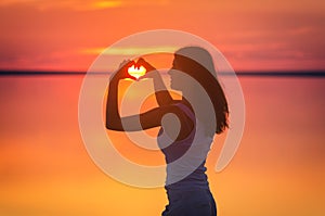 Beautiful female model enjoying sunset and making heart sign on sun. Calm water of salt lake Elton reflects woman`s silhouette. Gi