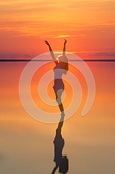 Beautiful female model open arms under sunset at seaside. Calm water of salt lake Elton reflects woman silhouette. Sun goes behind