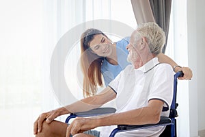 Beautiful female nurse taking care of senior patient in wheelchair at home, disabled elderly man sitting in wheelchair, young