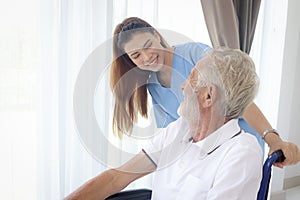 Beautiful female nurse taking care of senior patient in wheelchair at home, disabled elderly man sitting in wheelchair, young