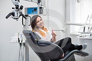 Beautiful female patient with perfect white teeth sitting in dental chair, smiling and showing thumbs up after treatment