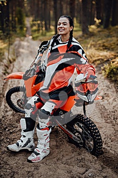 Beautiful female racer wearing protective motocross outfit smiling and looking on the camera while sitting on her bike