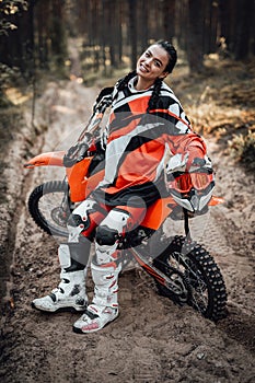 Beautiful female racer wearing protective motocross outfit smiling and looking on the camera while sitting on her bike