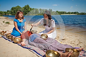 Beautiful female receiving energy sound massage with singing bowls and body massage on a river bank