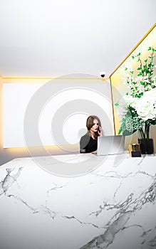 Beautiful female receptionist answering the phone and using laptop. Blank screen for mockup design
