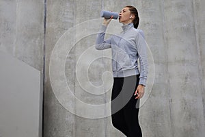 Beautiful female runner standing outdoors, drinking water from bottle. Fitness woman takes a break after running workout