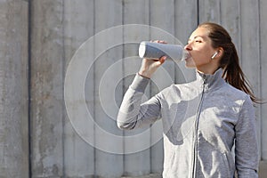 Beautiful female runner standing outdoors, drinking water from bottle. Fitness woman takes a break after running workout