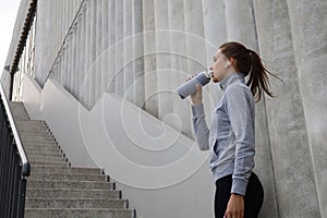 Beautiful female runner standing outdoors, drinking water from bottle. Fitness woman takes a break after running workout