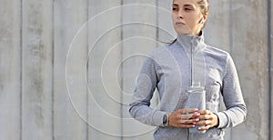 Beautiful female runner is standing outdoors holding water bottle. Fitness woman takes a break after running workout