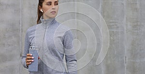 Beautiful female runner is standing outdoors holding water bottle. Fitness woman takes a break after running workout