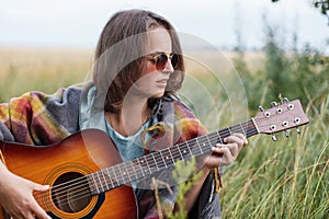 Beautiful female with short hairstyle wearing stylish sunglasses resting outdoors playing guitar enjoying her summer vacations. Fe