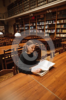 Beautiful female student studying in the library in the reading room, reading a book on the background of shelves with books.