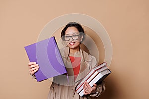 Beautiful female teacher educator professor in eyeglasses, smiling at camera while holding out book on beige background
