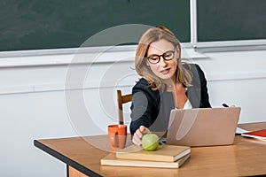 Beautiful female teacher in glasses sitting at computer desk and reaching for apple