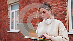 Beautiful female with wireless headphones on neck typing on laptop outdoors