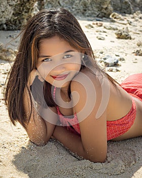 Beautiful Filipina in a red swimsuit smiling and resting head on a hand lying on the beach sand