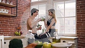 Beautiful fit couple make clicking with glass bottles drinking healthy cocktail after workout, talking and laughing in