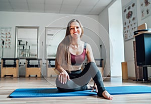 Beautiful fit-shape female dressed sporty sitting and sincerely smiling on sporty gym mat in sport athletic gym. Active people