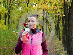 Beautiful fitness sport girl in autumn park in sportswear drinks water or isotonic drink from a sports bottle.