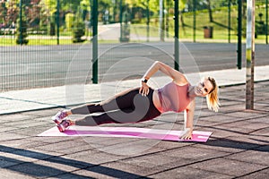 Beautiful fitness woman in tight pants training on mat outdoor summer day, doing side plank pose with one hand