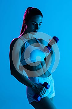 Beautiful fitness woman working out with dumbbells over blue light background