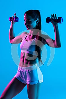 Beautiful fitness woman working out with dumbbells over blue light background