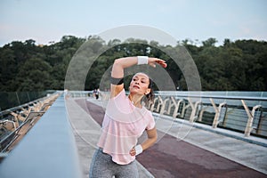 Beautiful fitness young woman exercising outdoors on the bridge, stretching her body before morning routine jog at sunrise. Sport