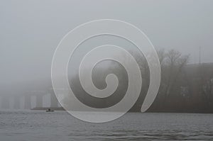 Beautiful foggy morning cityscape. Paton Bridge in rich fog, mist background. A man is fishing on an inflatable boat.