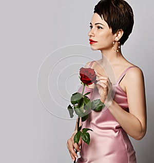 Beautiful fragile and tender brunette woman with short hair in pink silk dress and earrings with red rose in her arms