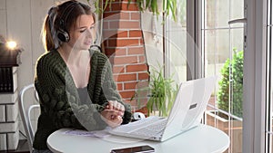 A beautiful freelance woman works on the line with a headset with a microphone and looks into a laptop webcam on the