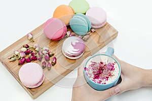 Beautiful french macarons on the desk. Woman hands holding blue cup of cappuccino with roses petals