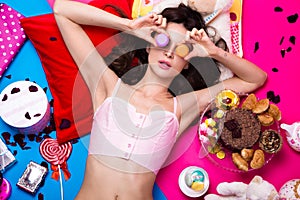 Beautiful fresh girl doll lying on bright backgrounds surrounded by sweets, cosmetics and gifts. Fashion beauty style.