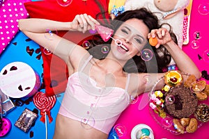 Beautiful fresh girl doll with soap bubbles lying on bright backgrounds surrounded by sweets, cosmetics and gifts