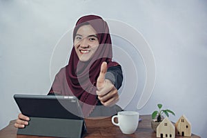 A beautiful, friendly young Muslim business woman smile and thumbs up at camera in hijab dress and suit using laptop