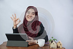 A beautiful, friendly young Muslim business woman smile and thumbs up okay at camera in hijab dress and suit using laptop