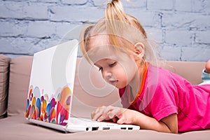 Beautiful funny blonde girl a child of two years is lying on the couch indoors and uses a white laptop computer technology with co