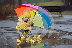 Beautiful funny blonde toddler boy with rubber ducks and colorful umbrella, jumping in puddles and playing in the rain