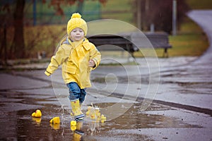 Beautiful funny blonde toddler boy with rubber ducks and colorful umbrella, jumping in puddles and playing in the rain