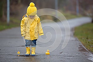 Beautiful funny blonde toddler boy with rubber ducks and colorful umbrella, jumping in puddles and playing in the rain