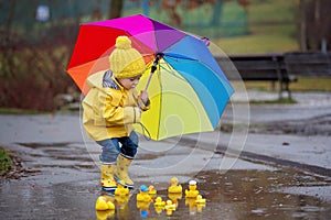 Beautiful funny blonde toddler boy with rubber ducks and colorful umbrella, jumping in puddles and playing in the rain