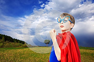 Beautiful funny child superhero wearing a cape standing in a green summer field