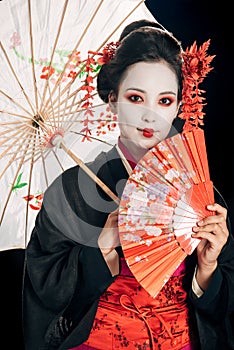Beautiful geisha in black kimono with red flowers in hair holding traditional asian umbrella and hand fan isolated on